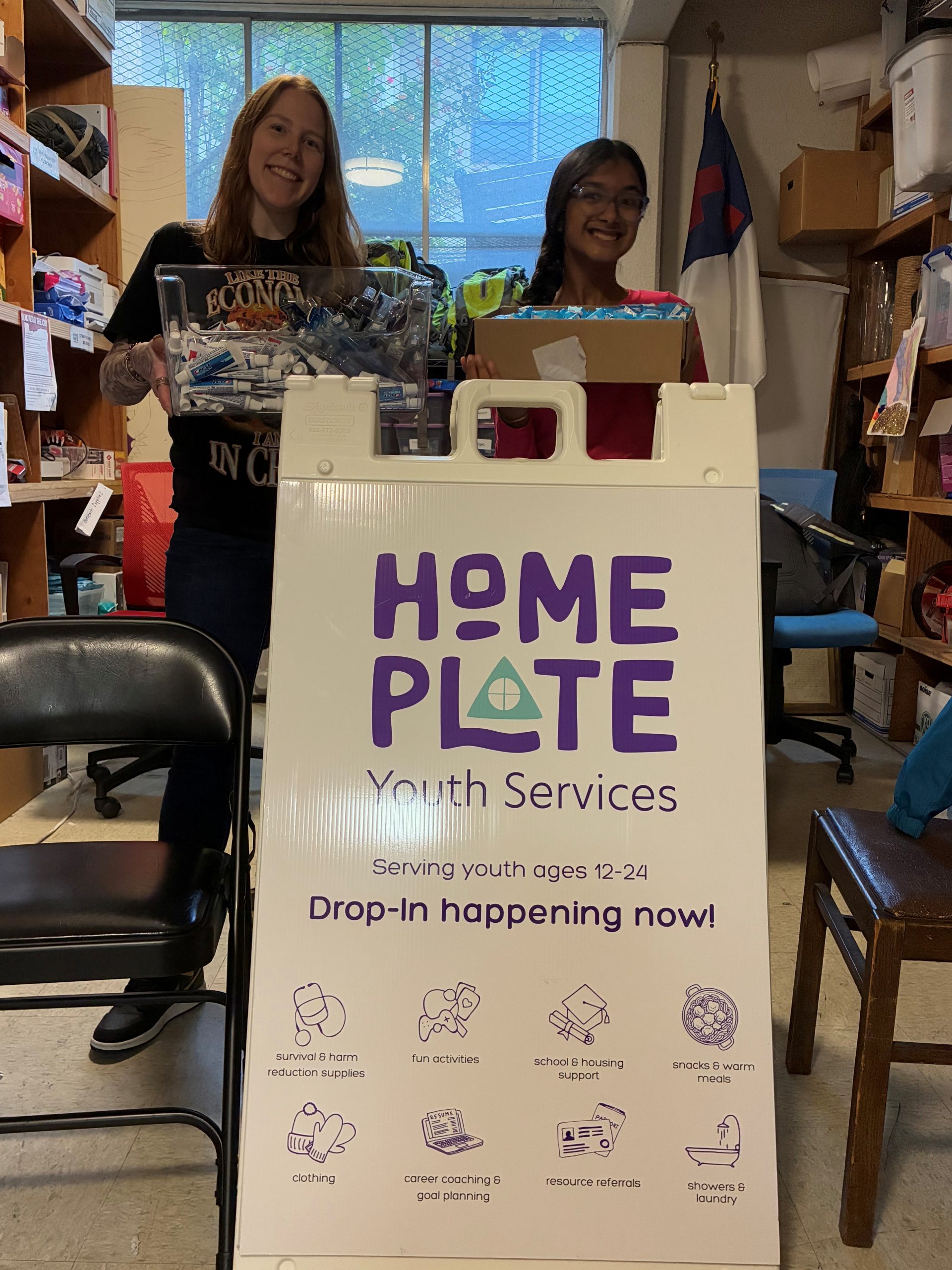 Saanvi and another woman stand in front of a sign for Home Plate Youth services holding containers of donated dental supplies.