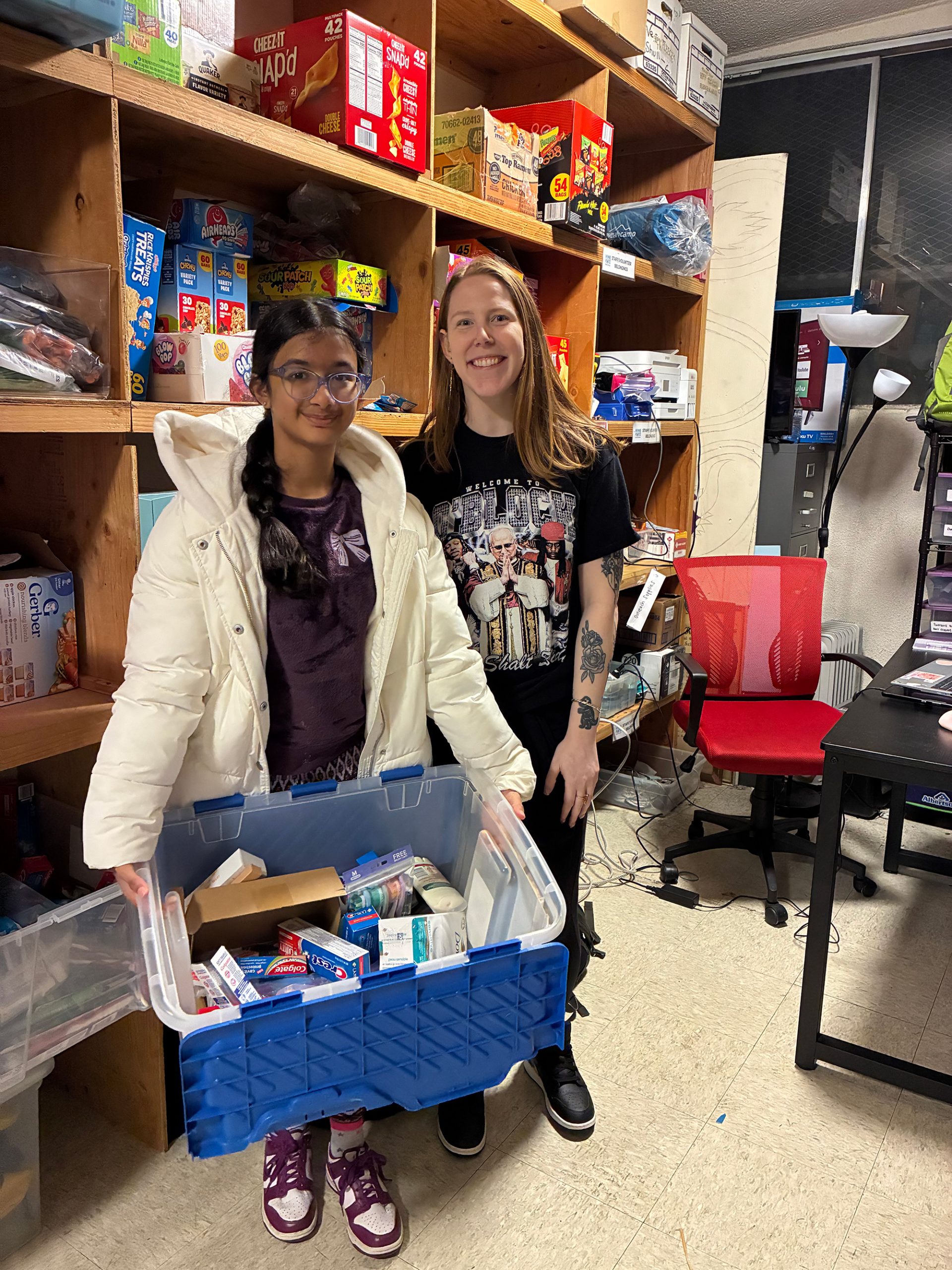 Saanvi stands next to another girl and his holding a large bin full of supplies for homeless like toothpaste, soap, and toothbrushes.