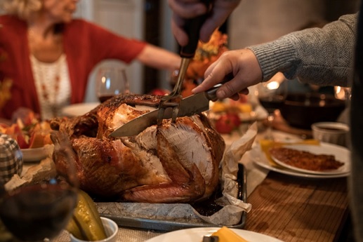 A roasted turkey on a table. A person with a fork & knife carves the turkey.